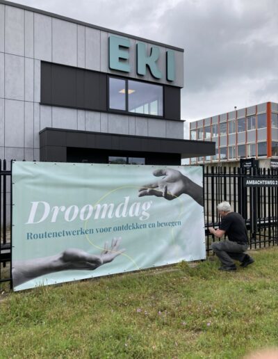 en man bevestigt een spandoek met de tekst “Droomdag – Routenetwerken voor ontdekken en bewegen” aan een hek bij de Papierfabriek in Nijmegen, als onderdeel van een creatieve sessie over toekomstbestendige routes.