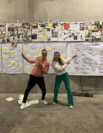 Michel Kolenbrander (links) en een collega presenteren ideeën en visies tijdens een creatieve sessie over routenetwerken, met posters en notities op een betonnen wand.