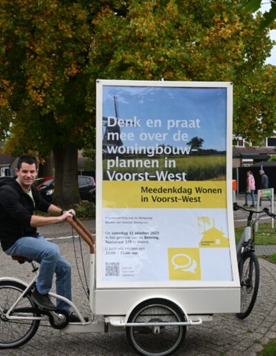 Man op een fietst met een groot mobiel promotiebord met informatie over woningbouwplannen in Voorst-West.